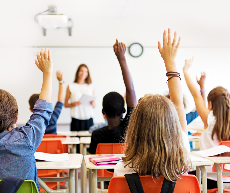 children in a classroom at school sitting at desks in rows facing the teacher with their hands up in the air